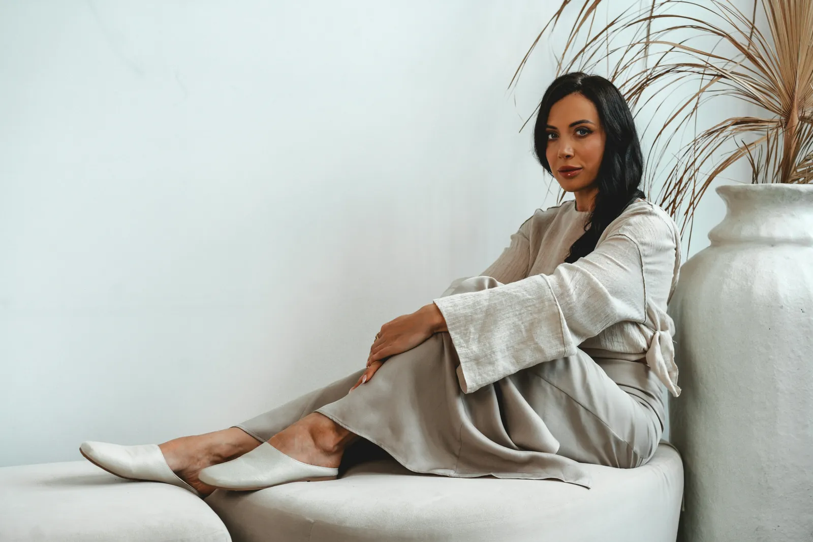 Woman sitting on a sofa in a relaxed linen top tied at the back and a draped satin midi skirt, a soft, unconstructed outfit